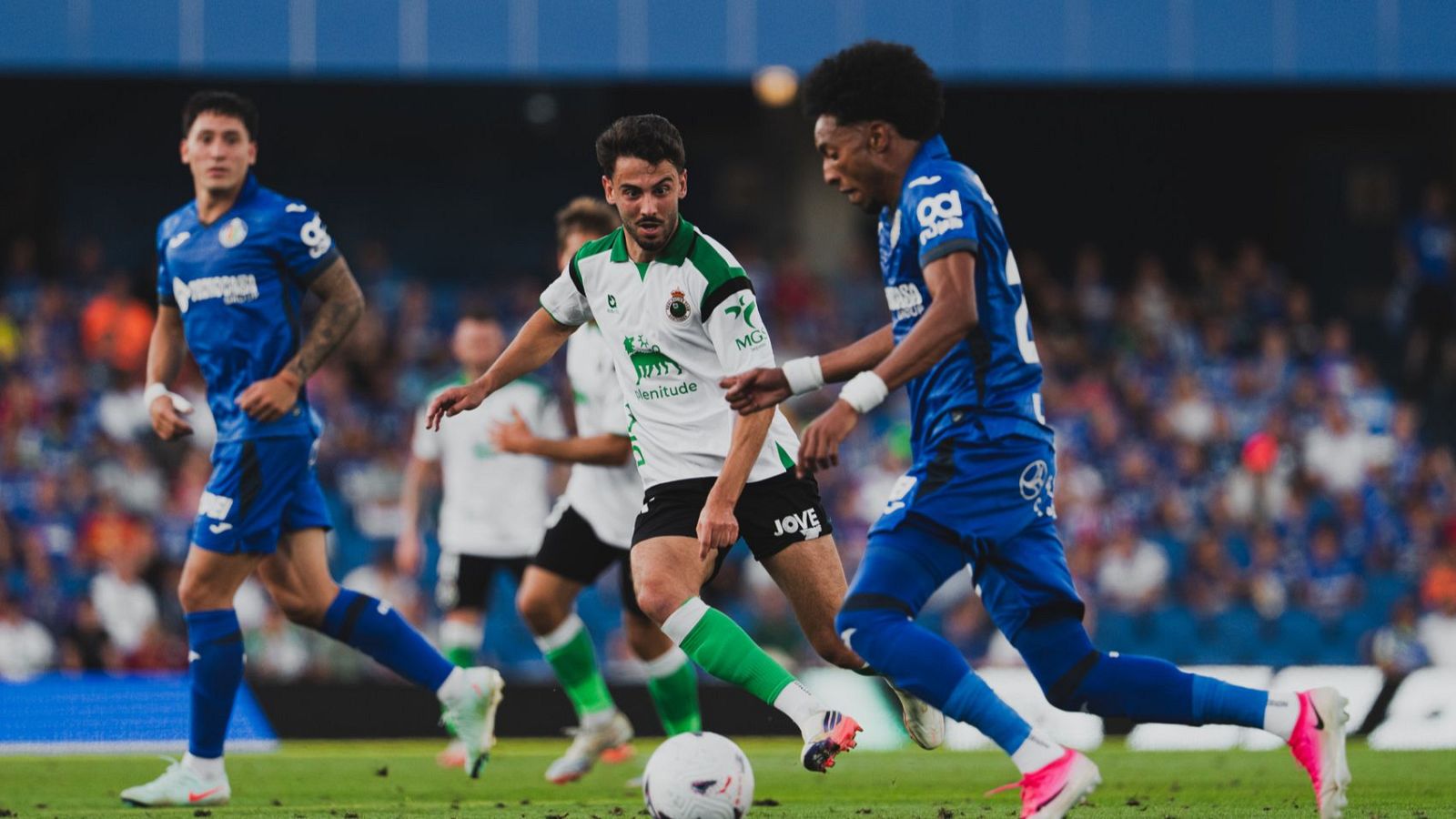 Jugador del Racing con balón es marcado por dos del Getafe CF en un partido. Se aprecian logos y dorsales en las camisetas. El fondo muestra gradas del estadio.