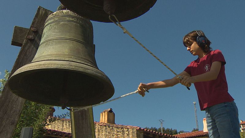 Joven con auriculares y camiseta roja haciendo sonar una campana de bronce en un entorno rural bajo un cielo azul.