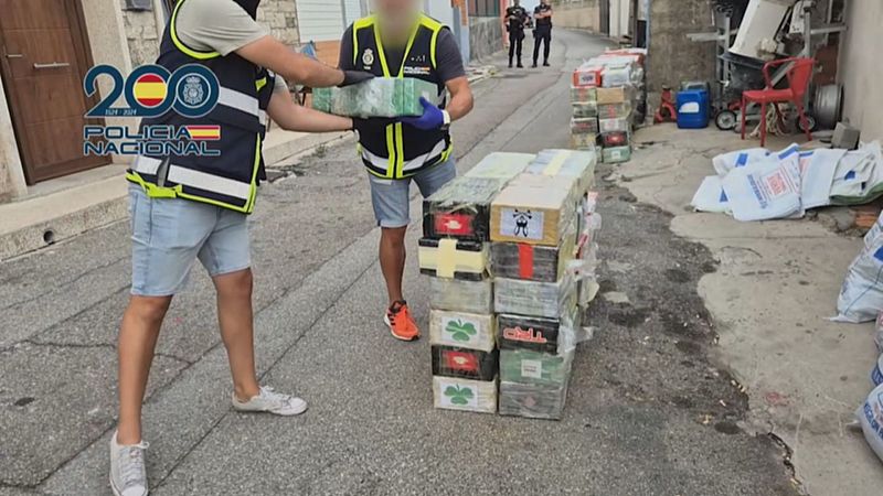 Axentes da Policía Nacional incautan gran cantidade de droga nunha rúa. Dous axentes manipulan paquetes envoltos en plástico, con marcas distintivas como cruces amarelas e debuxos de monos. O entorno é unha rúa urbana con muros e restos.
