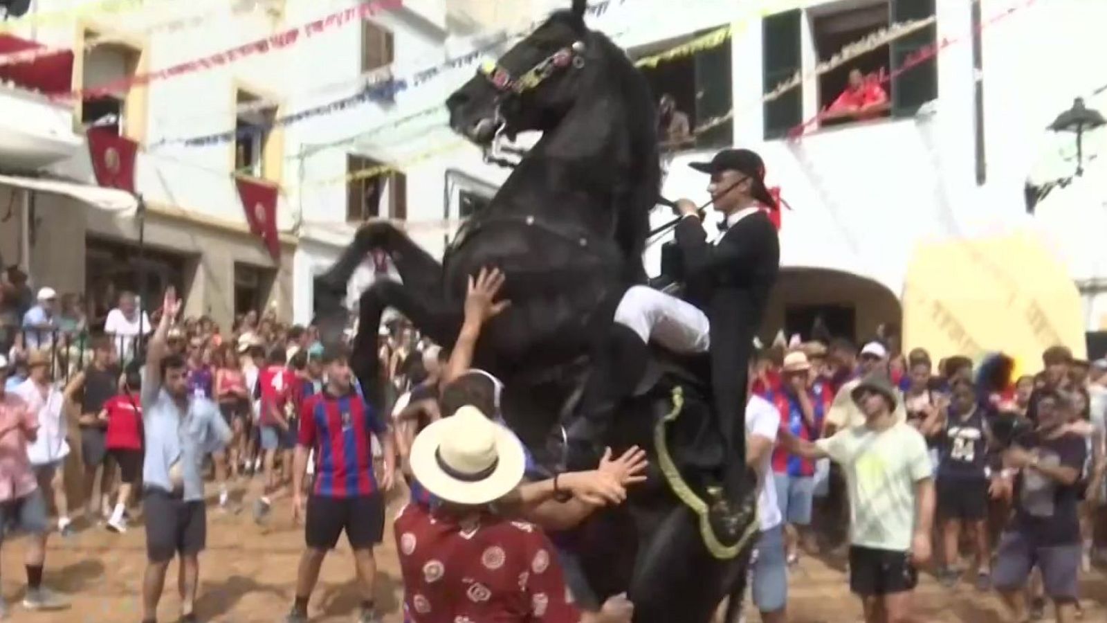 Caballo negro encabritado en el "Jaleo" de Ferreries, Menorca, con jinete y multitud interactuando. Edificios blancos y decoraciones festivas al fondo.