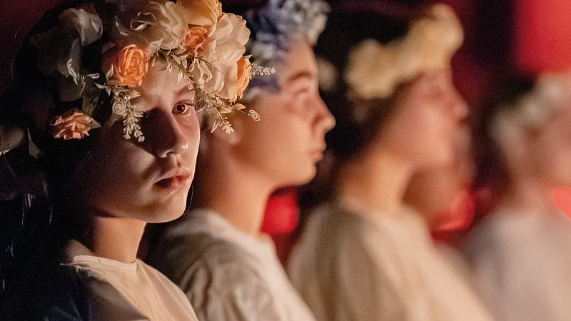 Jóvenes con coronas florales en fila, destacando una en primer plano con flores crema y melocotón, bajo una luz rojiza.