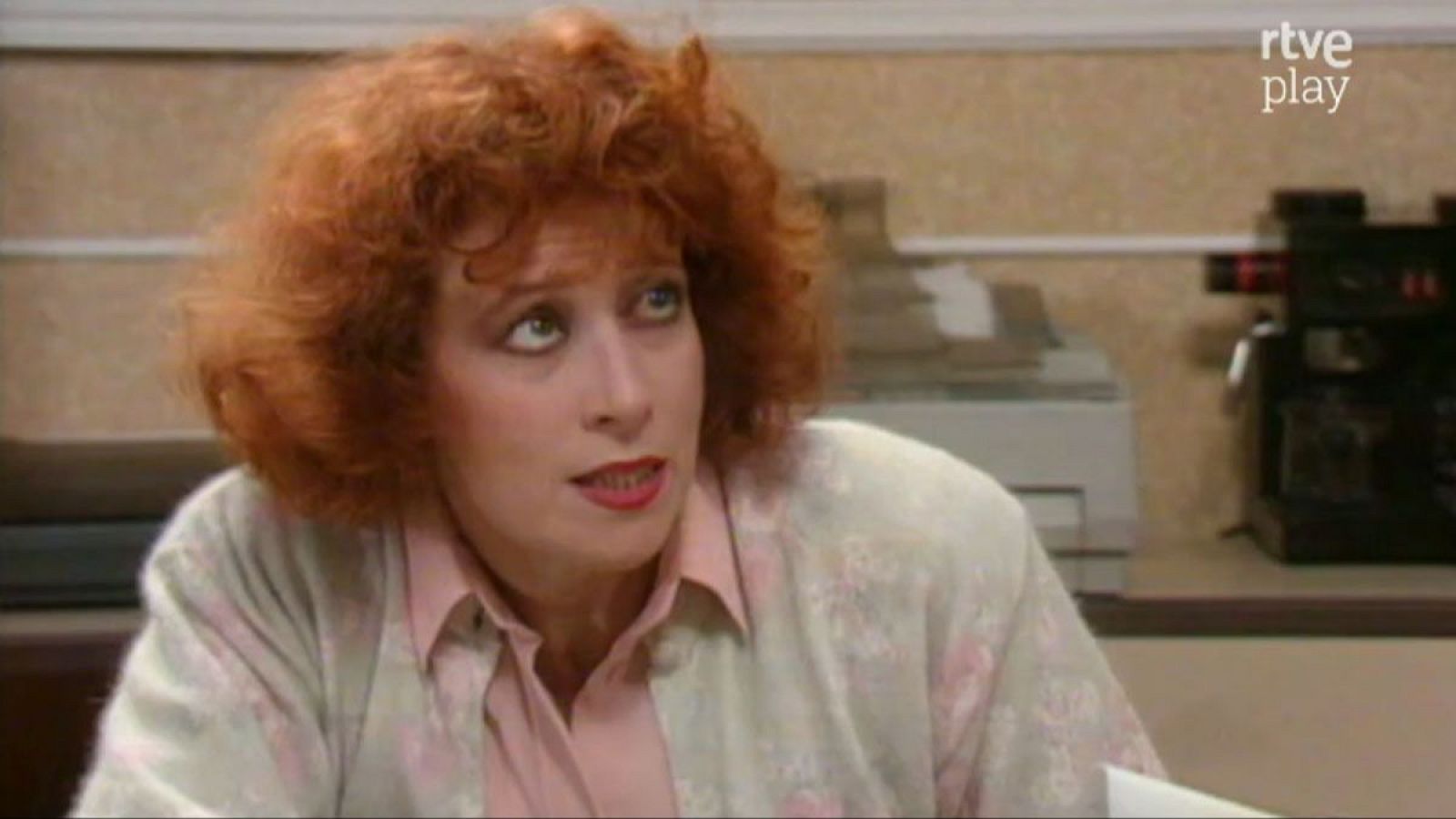 Primer plano de Mónica Randall, actriz española, con cabello pelirrojo rizado, ojos verdes y labios rojos. Expresión de sorpresa, boca abierta. Viste camisa rosa pálido y cárdigan claro. Fondo de oficina con máquina de café y logo "rtve play".