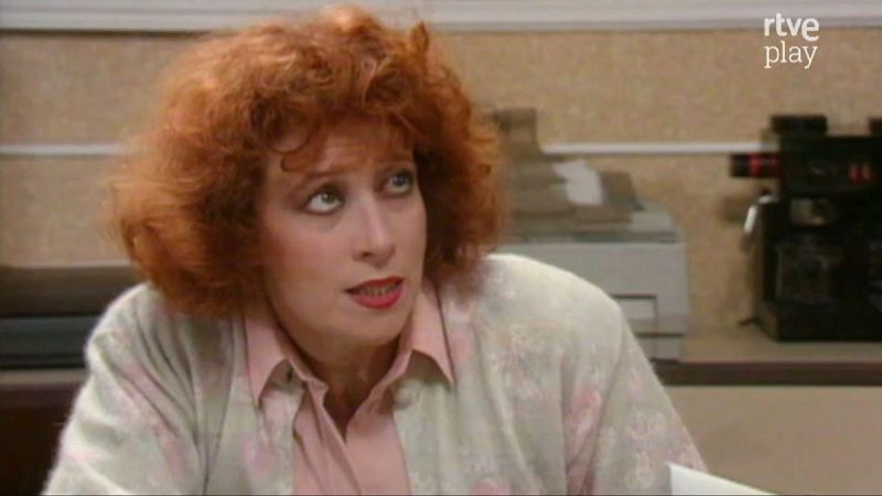 Primer plano de Mónica Randall, actriz española, con cabello pelirrojo rizado, ojos verdes y labios rojos. Expresión de sorpresa, boca abierta. Viste camisa rosa pálido y cárdigan claro. Fondo de oficina con máquina de café y logo "rtve play".
