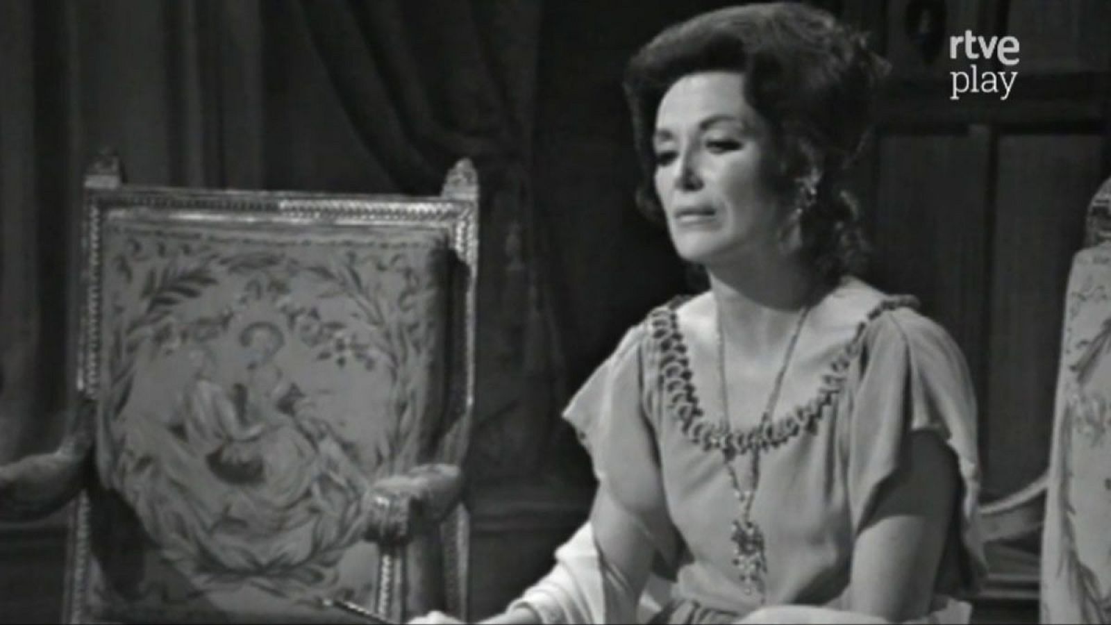 Primer plano de María Asquerino con peinado recogido, vestido y collar, junto a un sillón clásico. Logo de "rtve play" en la esquina superior derecha.