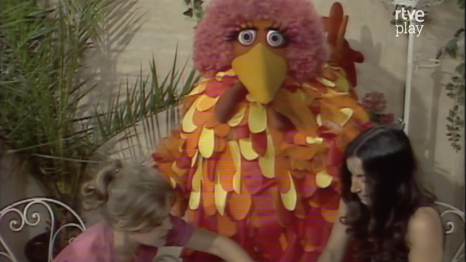 Caponata, el títere de "Barrio Sésamo" con cabello rosa y cuerpo de plumas, posa junto a dos mujeres en un escenario con vegetación. Se observa el logo "rtve play" en la esquina superior derecha.