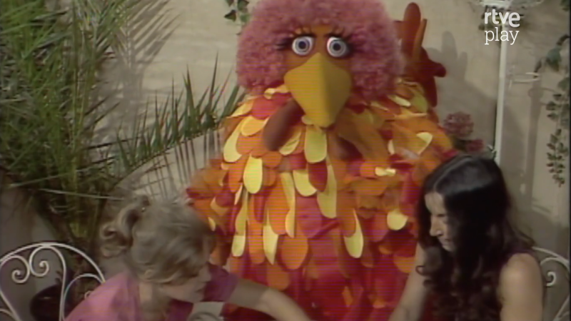 Caponata, el títere de "Barrio Sésamo" con cabello rosa y cuerpo de plumas, posa junto a dos mujeres en un escenario con vegetación. Se observa el logo "rtve play" en la esquina superior derecha.