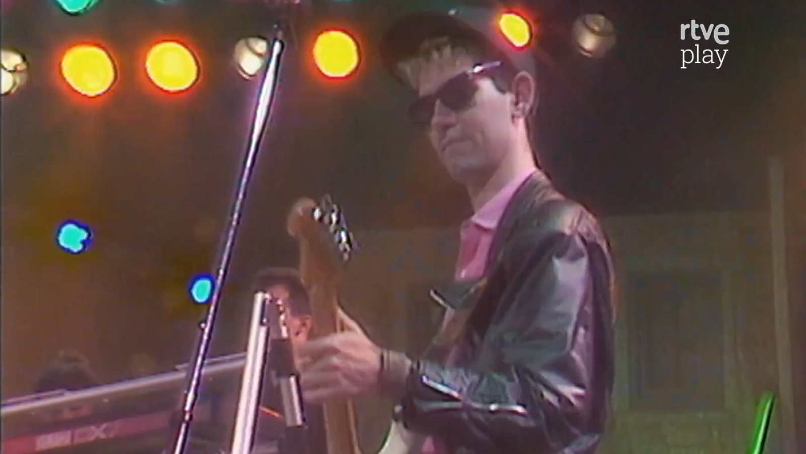 Carlos Berlanga, músico español, en concierto con gorra, gafas de sol, camisa rosa y chaqueta de cuero, tocando un instrumento de cuerda. Fondo con luces de escenario y logo "rtve play".