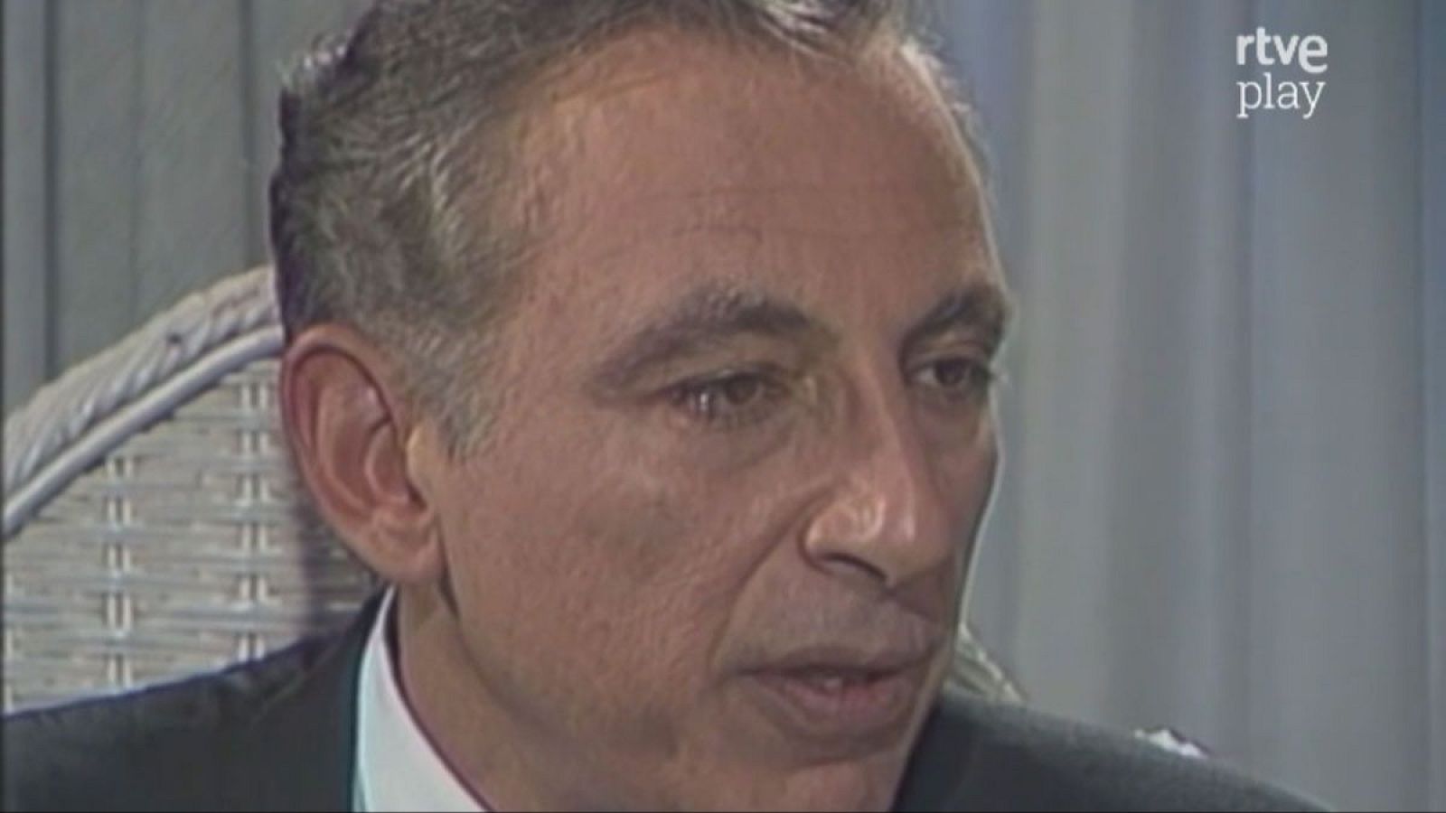 Robert Gallo, virólogo, de perfil con expresión seria. Viste traje oscuro y camisa blanca. Fondo desenfocado con logo "rtve play".