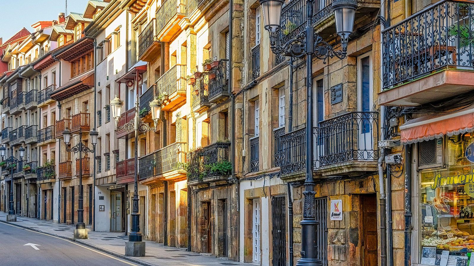Calle urbana en Oviedo con edificios tradicionales, balcones ornamentados y una frutería visible. Luz solar cálida y acera con patrón de baldosas.