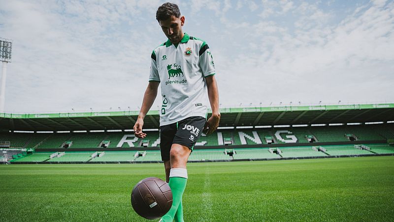 Iván Martín, futbolista español, en el césped de un estadio. Equipación blanca con detalles verdes y negros, patrocinadores 'plenitude' y 'JOVE GROUP'. Balón de fútbol clásico cerca.