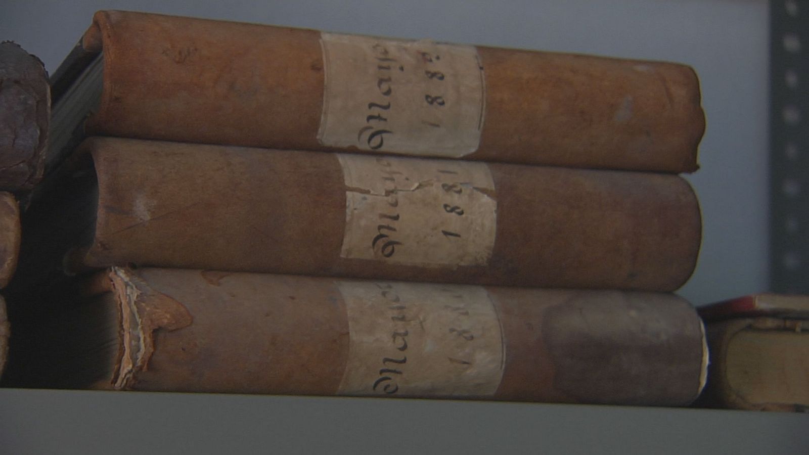 Tres volúmenes antiguos apilados, con etiquetas que muestran "1881" y "Manuscrito" escrito a mano. El estado de los libros indica su antigüedad.