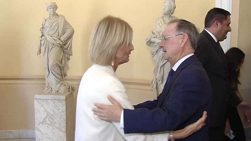 Isabel García Pelayo y Juan Vivas se abrazan en un interior formal, rodeados de estatuas clásicas. Ambos sonríen, mostrando un saludo cordial.