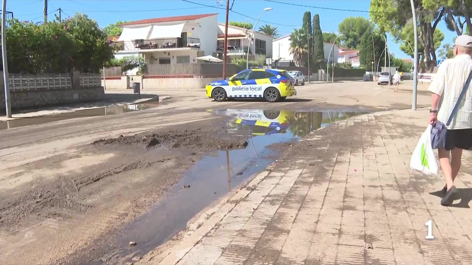 Coche de Policía Local en calle embarrada con charcos. Hombre mayor camina por acera con bolsa. Casas residenciales y vegetación al fondo. Se ve un '1' en la esquina inferior derecha.