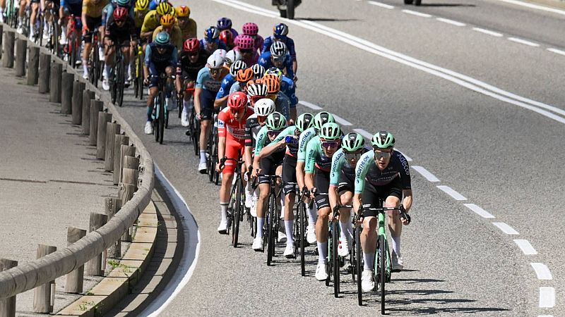 Peloton de ciclistas profesionales en carrera de carretera. Destacan ciclistas con maillots negro y verde claro con logos de "DECATHLON" y "adecco". Entorno natural y soleado.