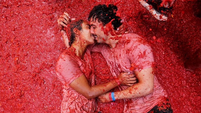 Un hombre y una mujer se besan intensamente, cubiertos de pulpa de tomate. Llevan camisetas manchadas y una pulsera de tela con franjas azules y rojas en la muñeca del hombre.