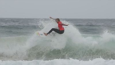 Surfista en acci�n sobre unha onda, con camiseta vermella e pantal�n escuro, sobre unha tabla branca. D�a nubrado na praia.