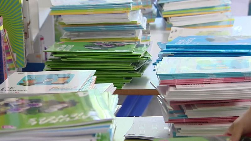 Superficie cubierta de pilas de libros y cuadernos con cubiertas verdes, azules y blancas. Se aprecian textos como "4.1 MATEMÁTICAS" y "SOCIAL SCIENCES". Una mano se vislumbra en la esquina inferior derecha.