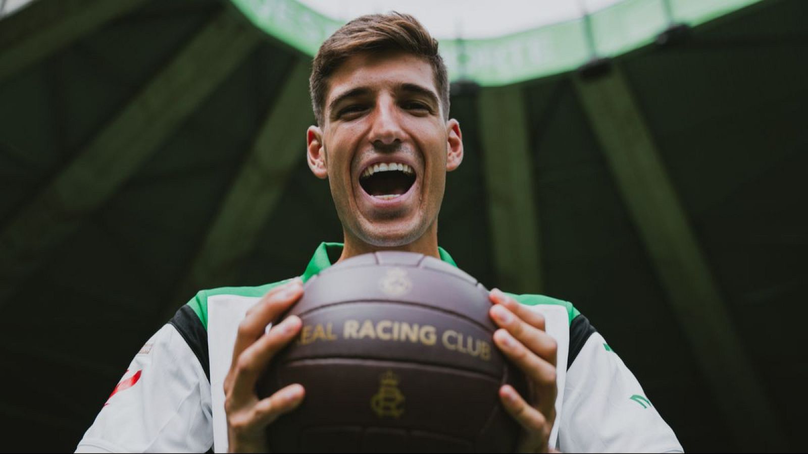 Matteo Prati sonríe con un balón del "REAL RACING CLUB" en sus manos, luciendo una camiseta deportiva blanca con franjas verdes. Fondo oscuro de estadio.