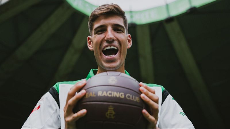 Matteo Prati sonríe con un balón del "REAL RACING CLUB" en sus manos, luciendo una camiseta deportiva blanca con franjas verdes. Fondo oscuro de estadio.