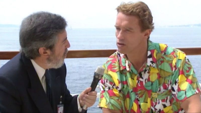 Arnold Schwarzenegger, con camisa tropical, es entrevistado por un hombre con traje oscuro frente a un mar azul y cielo despejado.