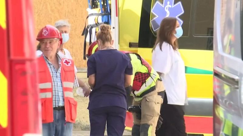 Emergencia al aire libre con personal de Cruz Roja, sanitario y de rescate. Voluntario de Cruz Roja en primer plano, ambulancia amarilla y verde visible. Personas con mascarillas atendiendo una situación.