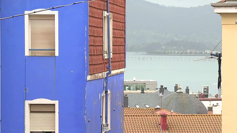 Vista urbana y costera de Santander con edificios de fachadas azul y marrón rojizo, tejados de terracota y un tejado abovedado gris. Al fondo, una bahía y montañas verdes bajo un cielo brumoso.