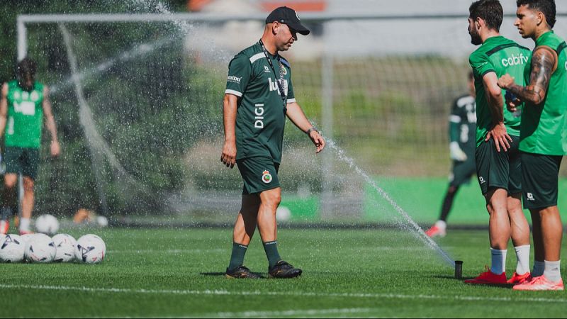 Entrenador de fútbol José Alberto López Menéndez, con uniforme "DIGI", en un campo verde con jugadores y balones. Aspersor activo.
