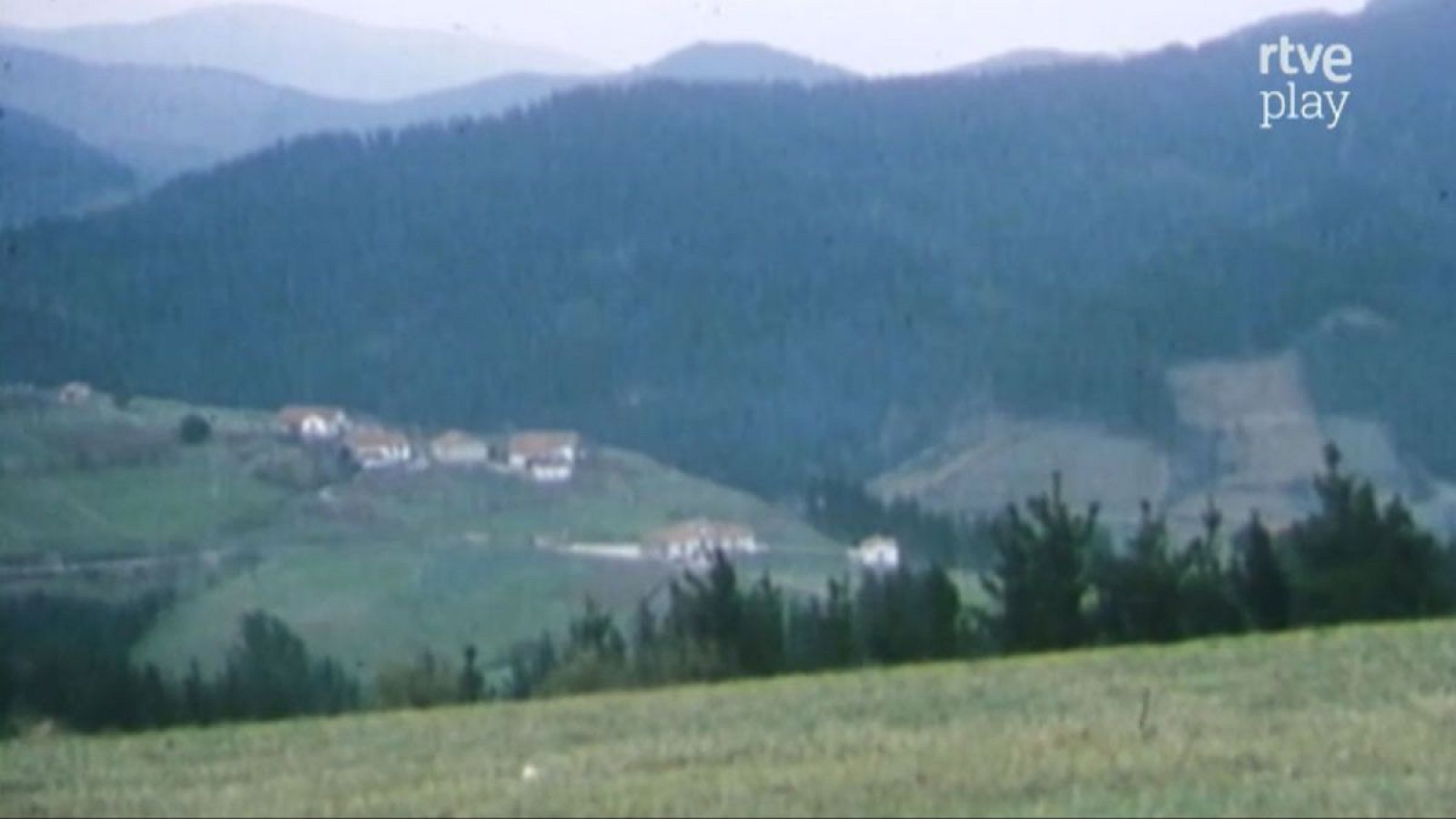 Paisaje montañoso y rural con pradera, caseríos dispersos y cadenas montañosas boscosas al fondo. Logotipo de "rtve play" en la esquina superior derecha.