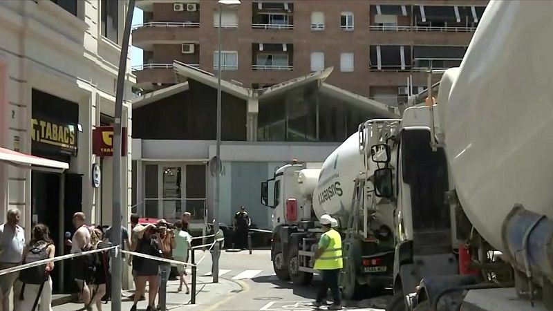 Obras en la calle con camiones hormigonera "Sustem" y cinta de balizamiento. En la acera, un "TABACS ESTANCO" y gente observando.