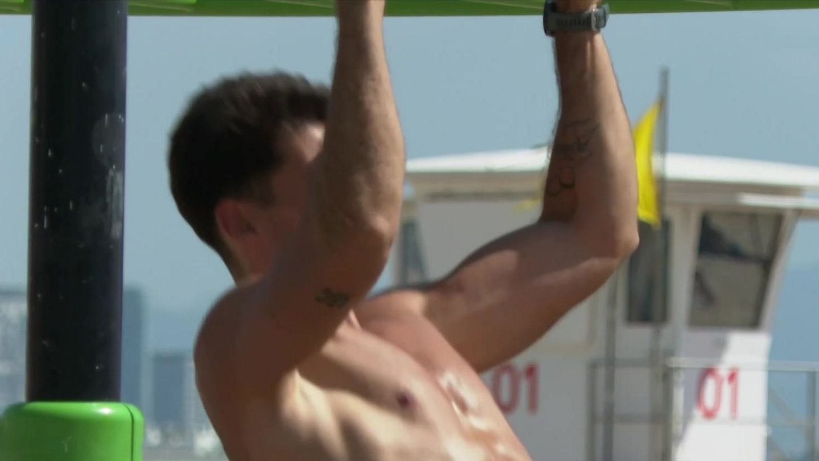 Hombre musculoso ejercitándose al aire libre, suspendido de una barra. Tatuajes visibles: "307" en un brazo, abstracto en el otro. Reloj deportivo. Torre de socorristas con "01".