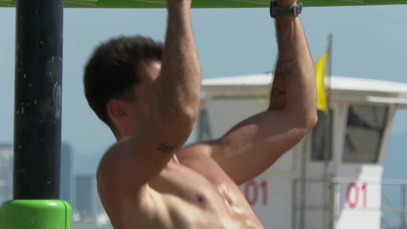 Hombre musculoso ejercitándose al aire libre, suspendido de una barra. Tatuajes visibles: "307" en un brazo, abstracto en el otro. Reloj deportivo. Torre de socorristas con "01".