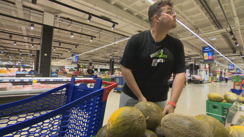 Hombre con gafas y camiseta "OPERACIÓN DESPENSA" mira melones en un supermercado. Fondo con estanterías y carro azul.