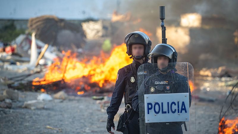 Agentes de la Policía Nacional con escudos y cascos antidisturbios, frente a un gran incendio y escombros en un entorno exterior.