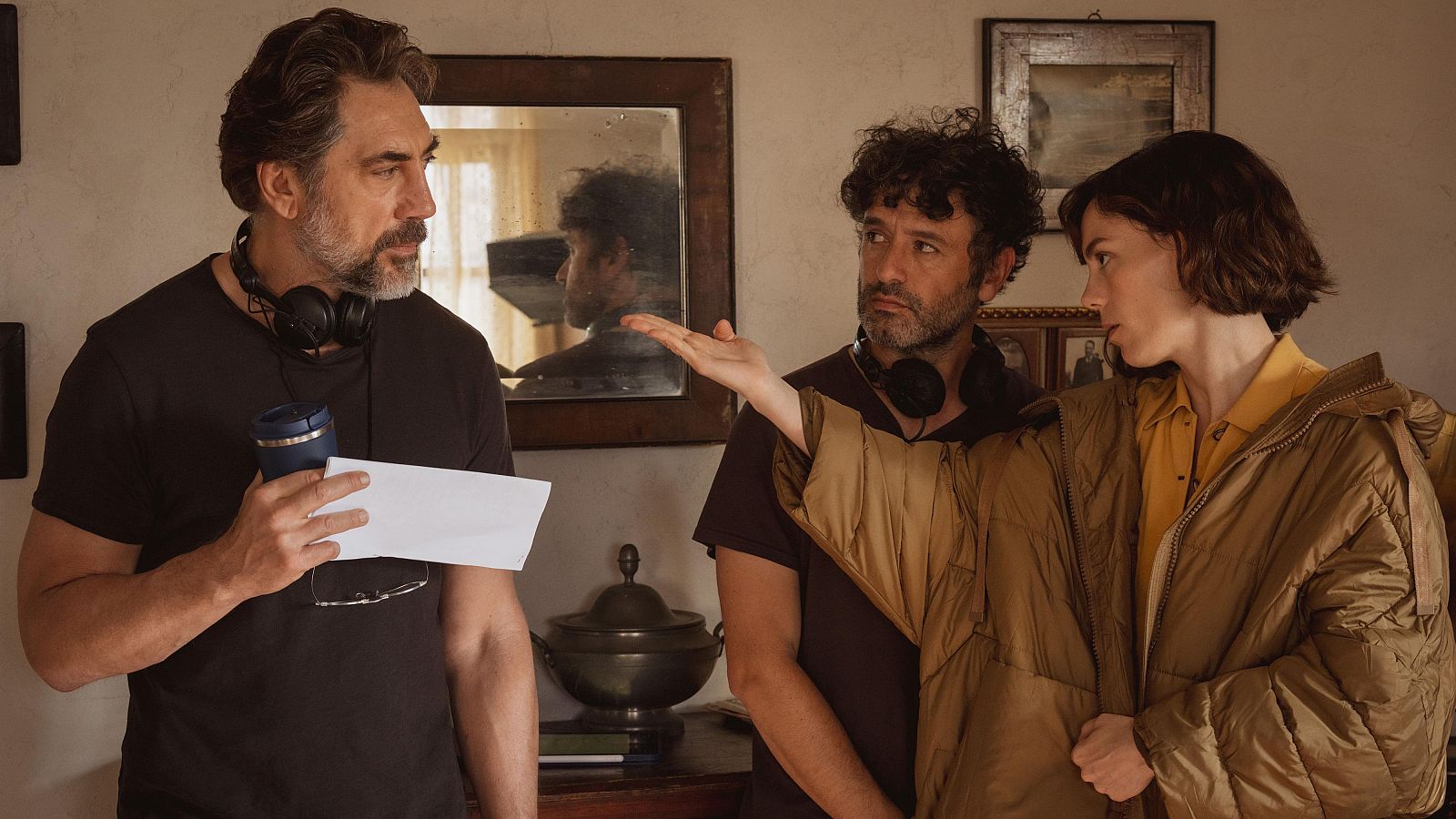 Javier Bardem, Fernando León de Aranoa y una mujer joven en una escena de producción. Bardem con papeles y objeto azul, Aranoa gesticula, la mujer escucha atentamente.