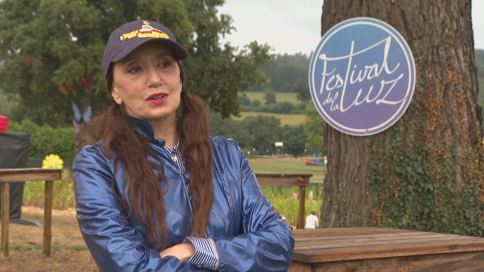 Luz Casal, con gorra y chaqueta azul, en el "Festival de la LUZ" en un entorno natural. Detrás, un cartel anuncia el evento.