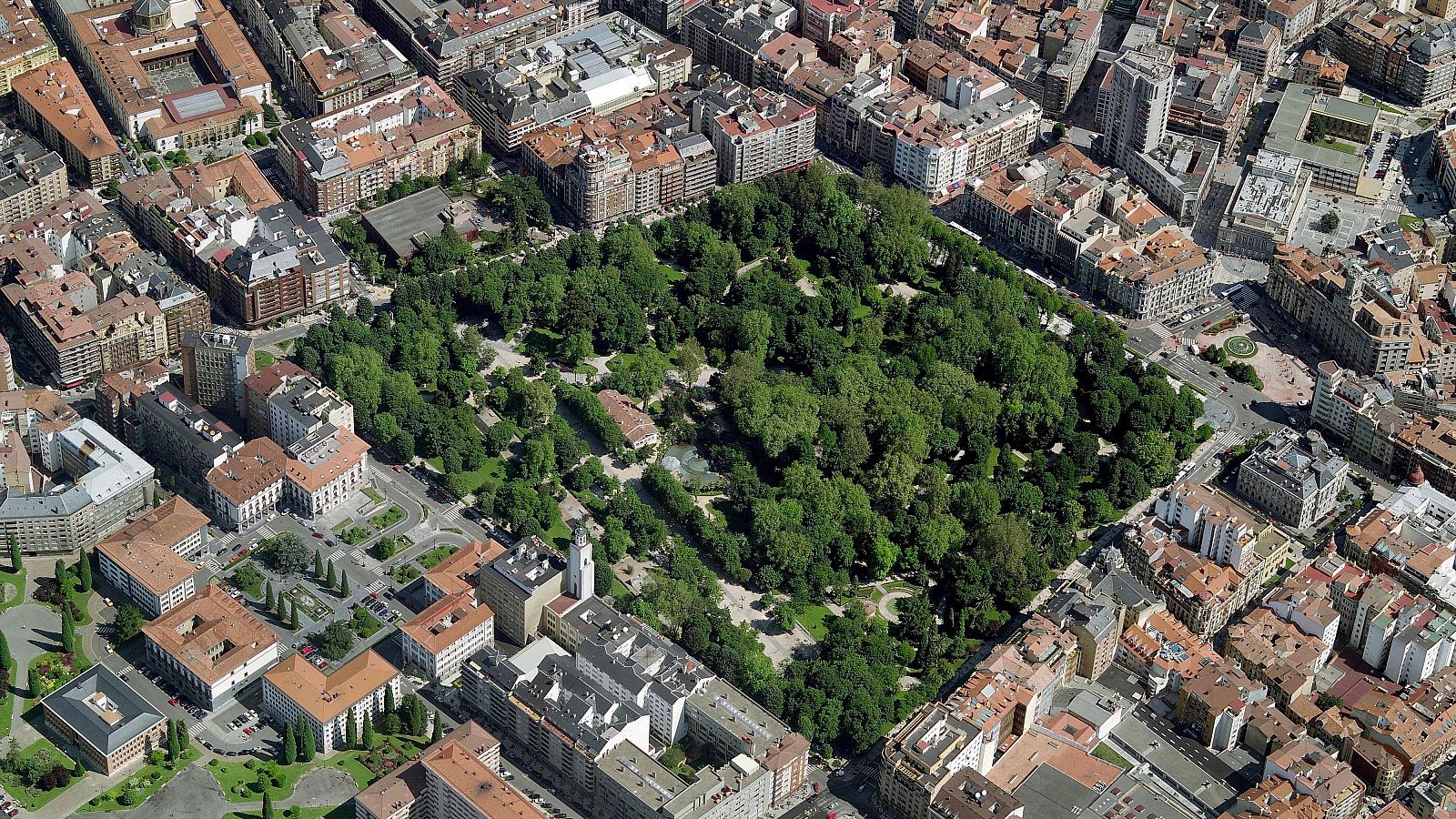 Vista cenital del Campo San Francisco, un parque urbano en Oviedo, Asturias, rodeado de edificios y calles. Se aprecia la vegetación frondosa y la arquitectura urbana.
