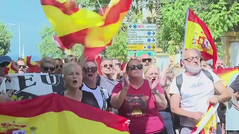 Concentración de personas en Ceuta ondeando banderas de España, con carteles y señales de tráfico visibles. Ambiente festivo o de manifestación.