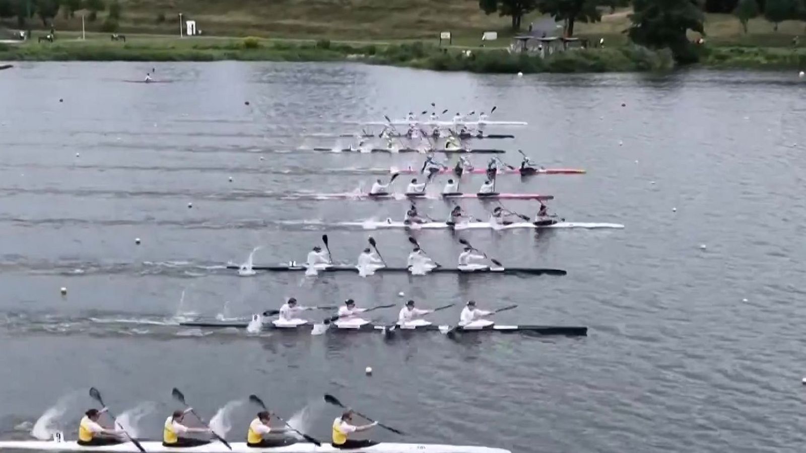 Vista aérea de una regata de kayaks K4 en plena competición, con embarcaciones en carriles delimitados por boyas y atletas remando intensamente. El agua está llena de salpicaduras y estelas.