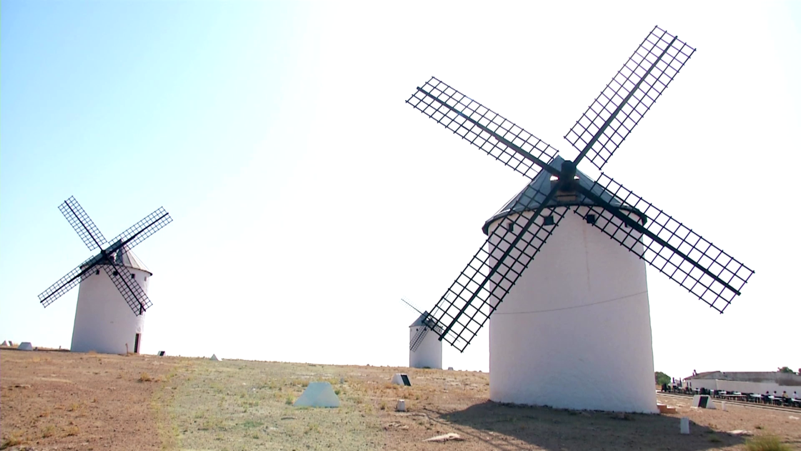 Tres molinos de viento blancos con aspas oscuras se alzan sobre una loma árida bajo un cielo azul brillante. El molino principal, en primer plano, es el más grande y detallado.