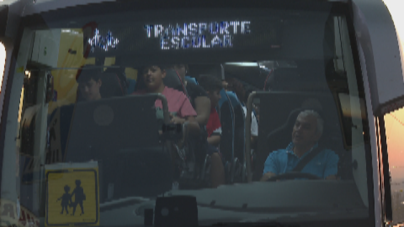 Autobús de transporte escolar con conductor y pasajeros infantiles, señal de escolares en el parabrisas y texto "TRANSPORTE ESCOLAR" en la parte superior.