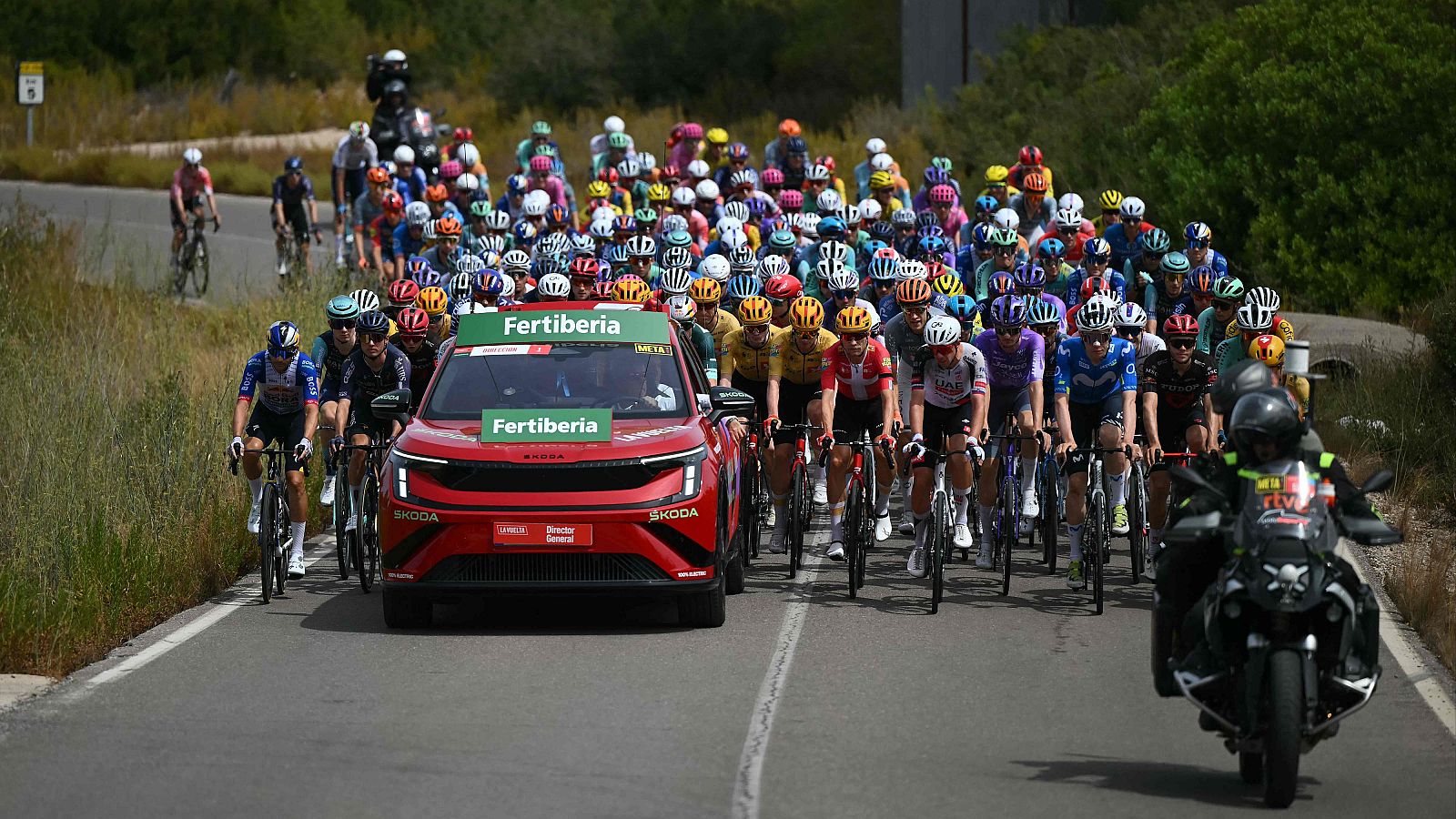 Pelotón ciclista profesional en carretera soleada, acompañado por coche "Fertiberia" (INEOS, META) y motocicleta. Señal "5" visible. Resumen Vuelta 7, Vuelta 2026.