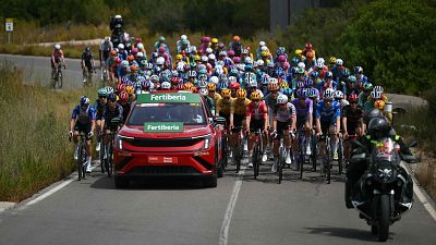 Pelot�n ciclista profesional en carretera soleada, acompa�ado por coche "Fertiberia" (INEOS, META) y motocicleta. Se�al "5" visible. Resumen Vuelta 7, Vuelta 2026.