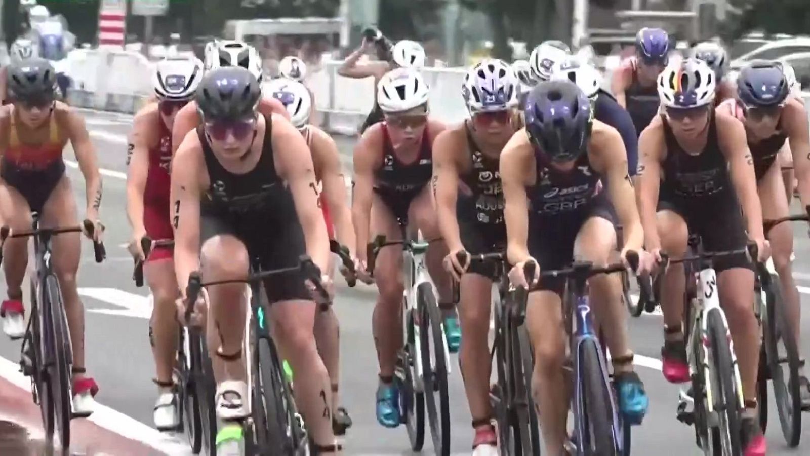 Grupo de ciclistas, predominantemente mujeres, compitiendo en un triatlón. Se aprecian cascos, gafas, trajes oscuros con "GBR" y dorsal "1" en una atleta. Bicicletas de carretera en una vía con barreras de seguridad.