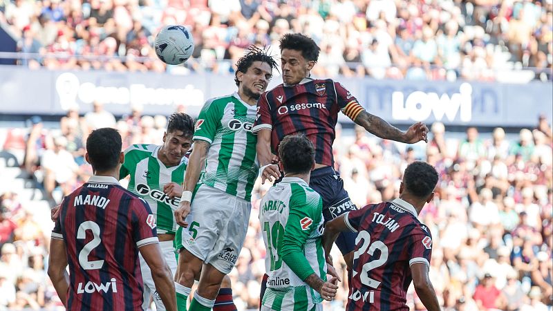 Jugadores Guido Rodríguez (Real Betis) y José Luis Morales (Levante UD) saltan por el balón en un partido de Primera División. Aficionados en las gradas y logotipos de patrocinadores visibles.