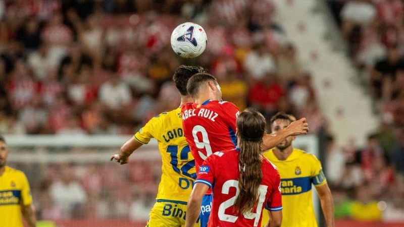 Jugadores compiten por un bal�n a�reo en un partido de f�tbol. Abel Ruiz (9, roja) y Loio (10, amarilla) son los protagonistas, con otros jugadores y el p�blico de fondo.