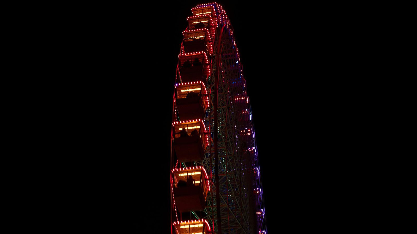 Noria iluminada con luces rojas, naranjas, púrpuras y azules contra fondo negro. Se aprecian cabinas y siluetas de personas. Fotografía nocturna desde ángulo bajo.