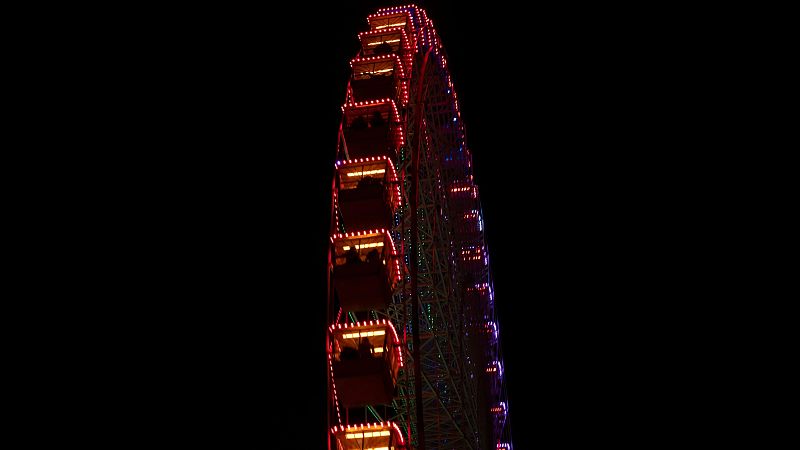 Noria iluminada con luces rojas, naranjas, púrpuras y azules contra fondo negro. Se aprecian cabinas y siluetas de personas. Fotografía nocturna desde ángulo bajo.