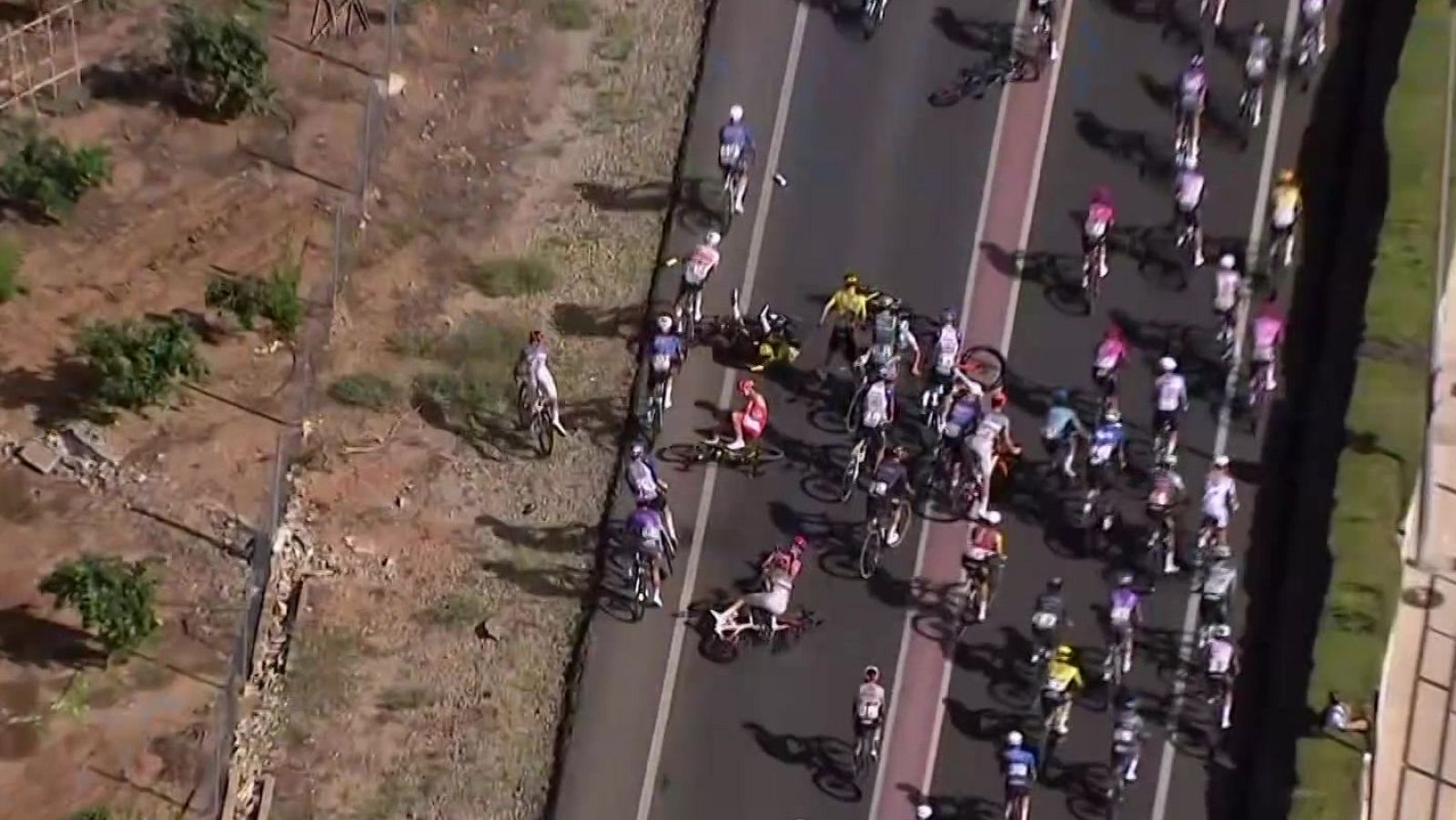 Caída masiva en carrera ciclista: ciclistas y bicicletas volcadas en la carretera. Varios corredores en el suelo, maillots de colores diversos.