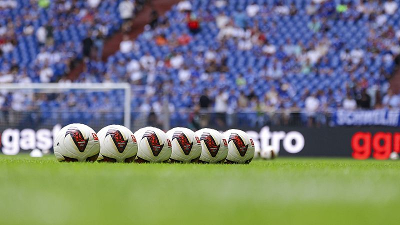 Balones de fútbol alineados en césped, con valla publicitaria "SCHWELM gg!" y gradas de estadio al fondo.