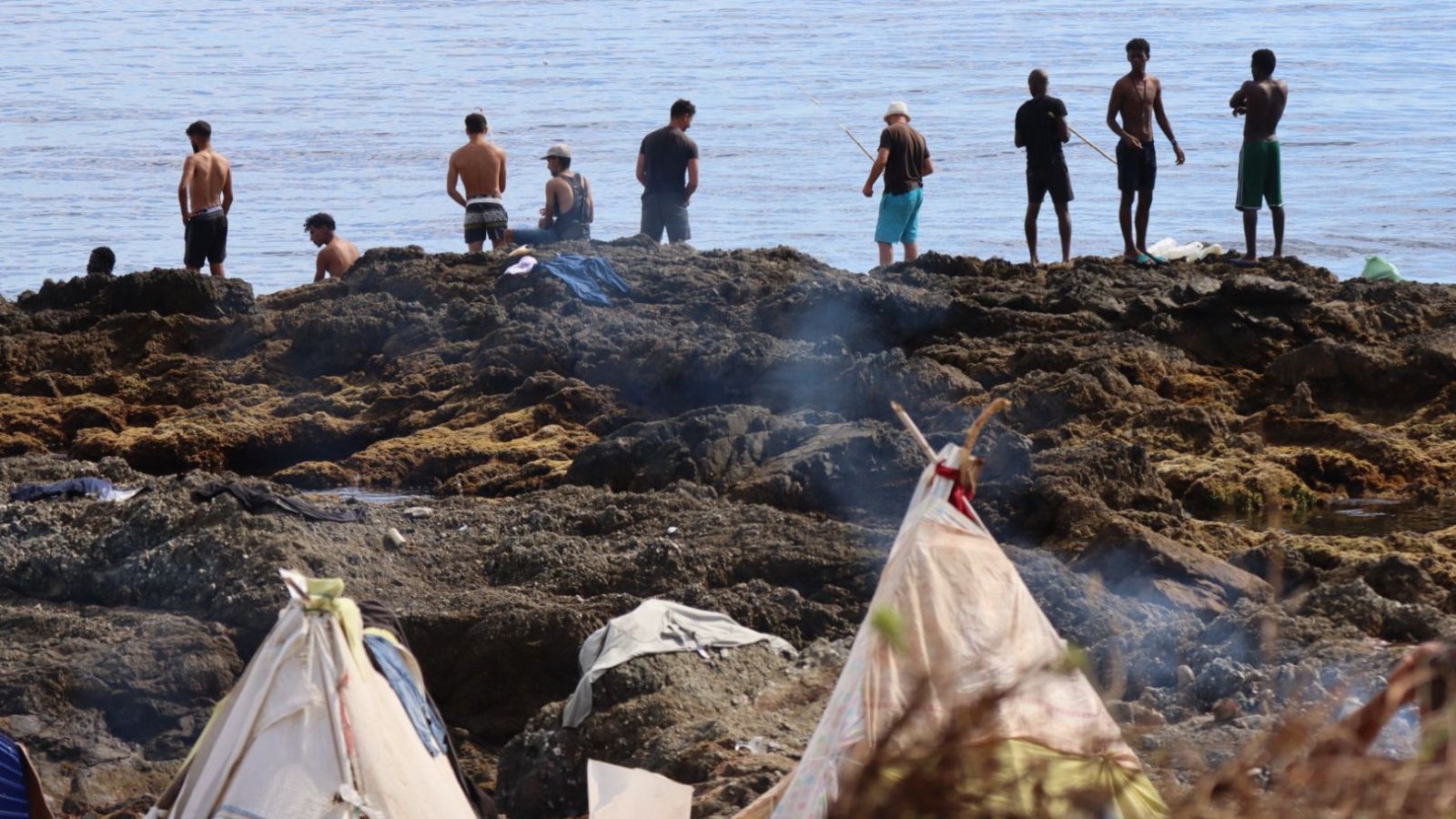 Costa rocosa con refugios improvisados, hogueras y nueve hombres, uno con caña de pescar, observando el mar. Fin de semana 24h.